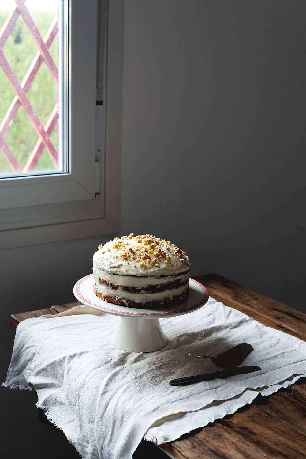 En una mesa de madera hay un mantel vintage blanco. Encima  Encima hay una portatarta blanca. Encima de ella está el Carrot Cake americano. 