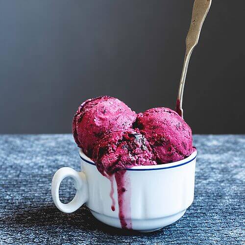 En una mesa hay un mantel gris. Encima hay una taza blanca llena de tres copas de helado de cerezas. Entre las bolas de helado hay una cuchara de té metida.