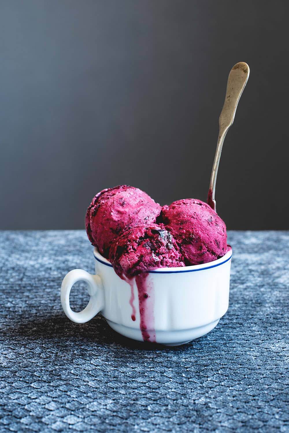 En una mesa hay un mantel gris. Encima hay una taza blanca llena de tres copas de helado de cerezas. Entre las bolas de helado hay una cuchara de té metida.