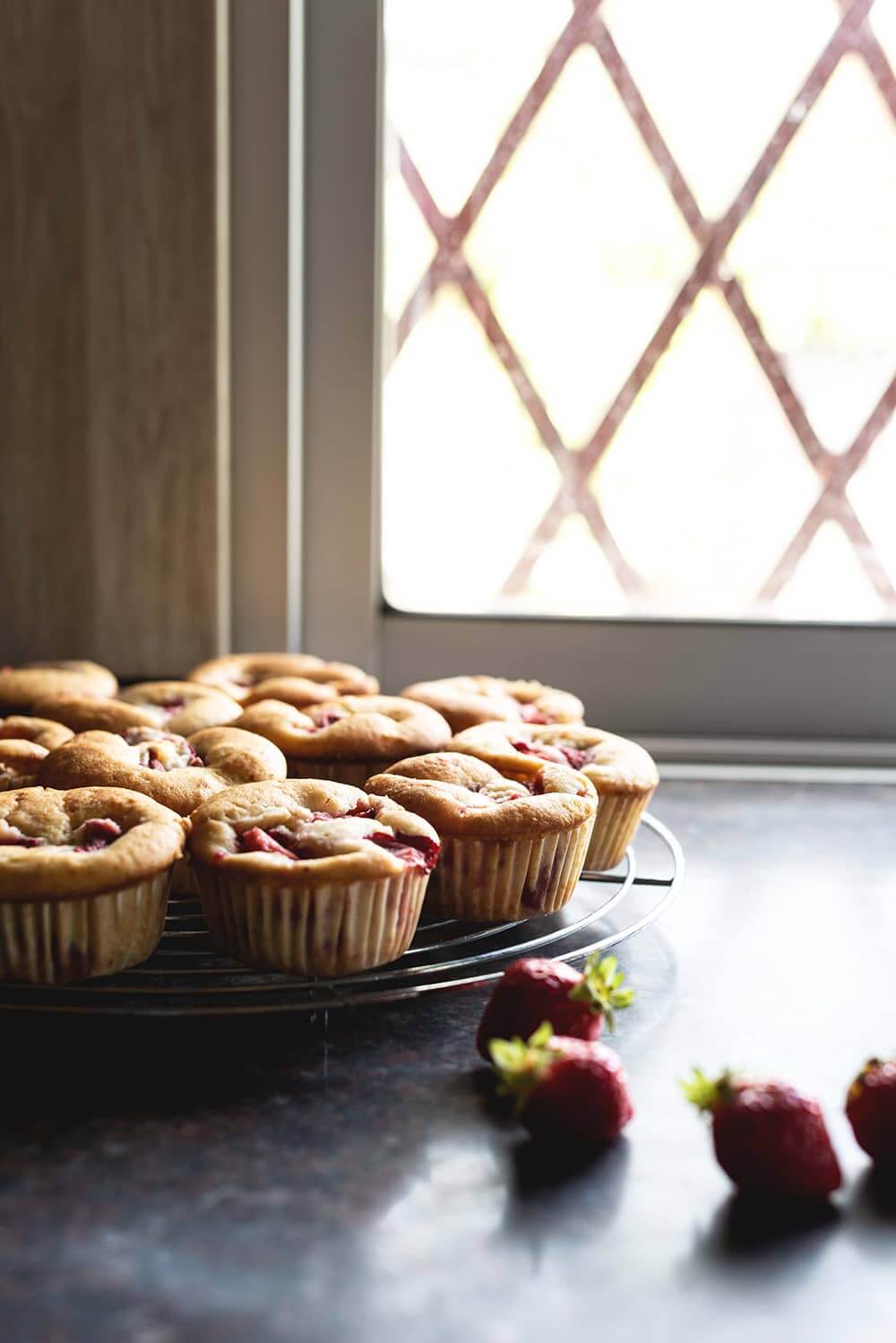Cómo hacer magdalenas con fresas En una encimera gris hay una rejilla. En la rejilla están las magdalenas con fresas. Alrededor de las magdalenas hay un par de fresas. Se ven bien cerca y suculentas.