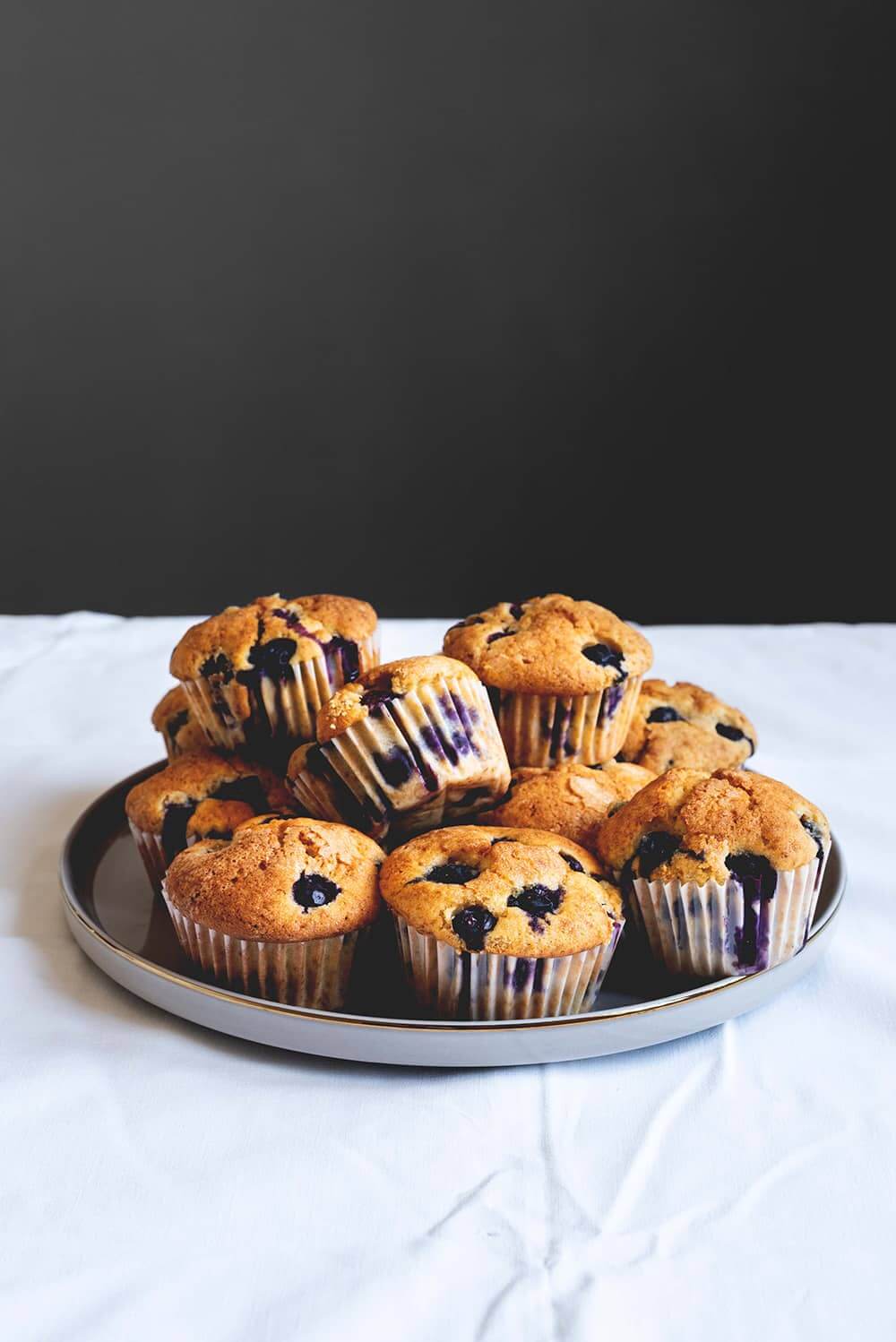En una mesa de madera hay un mantel blanco. Encima hay un plato gris encima están muchos muffins de arándanos. Se ven deliciosos y dorados.