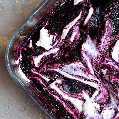 En una mesa gris hay un recipiente transparente lleno de helado de frutos rojos.Se ve marmolado donde se mezcla el sabor helado natural y dulce con helado del sabor de frutos rojos.