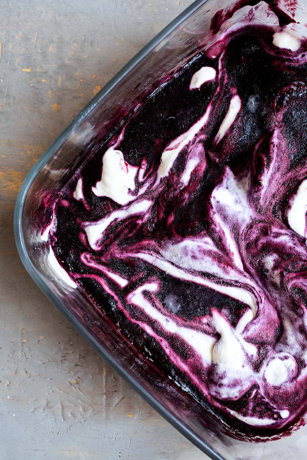 En una mesa gris hay un recipiente transparente lleno de helado de frutos rojos.Se ve marmolado donde se mezcla el sabor helado natural y dulce con helado del sabor de frutos rojos.