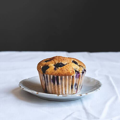 En una mesa de madera hay un mantel blanco. Encima hay un plato blanco encima está el muffin de arándanos. Se ven los pliegues de la cápsula del papel de magdalena. El postre se ve jugoso.