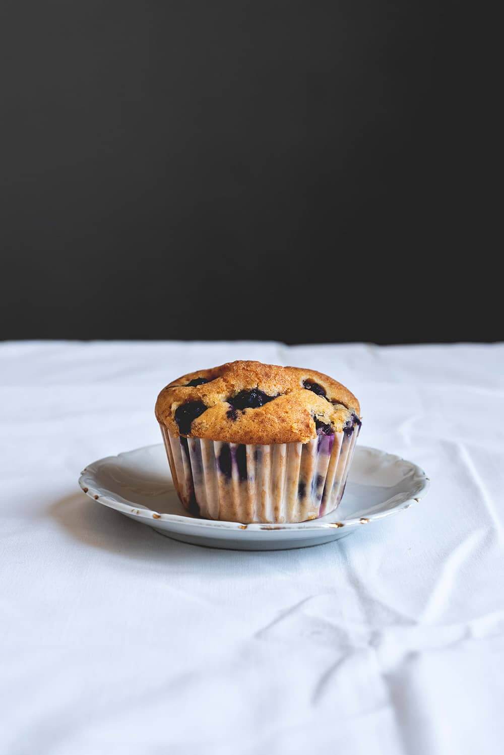 En una mesa de madera hay un mantel blanco. Encima hay un plato blanco encima está el muffin de arándanos. Se ven los pliegues de la cápsula del papel de magdalena. El postre se ve jugoso.