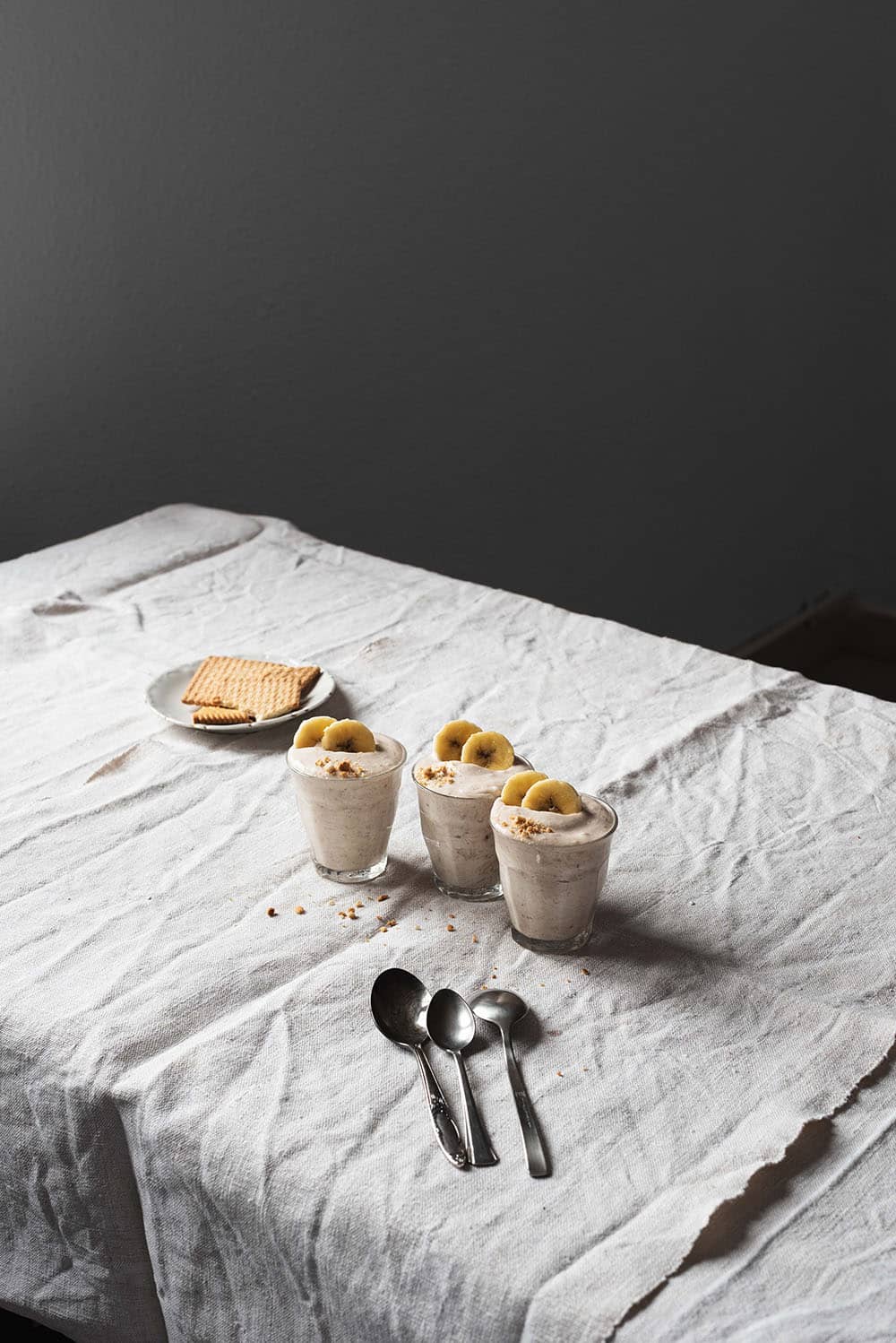 En una mesa hay un mantel blanco. Encima hay tres vasos transparentes de mousse de plátano. En un plato aparte galletas. También se ven cucharas de té y migas en la mesa.