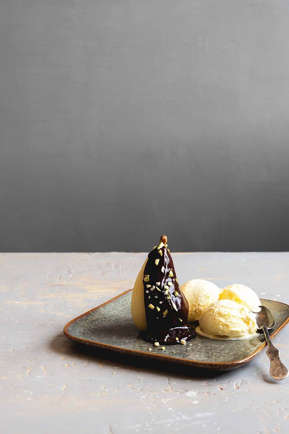 En una mesa gris hay un plato gris. Encima está el postre Peras al estilo helene. Al lado del las bolas de helado hay una cuchara de té. 