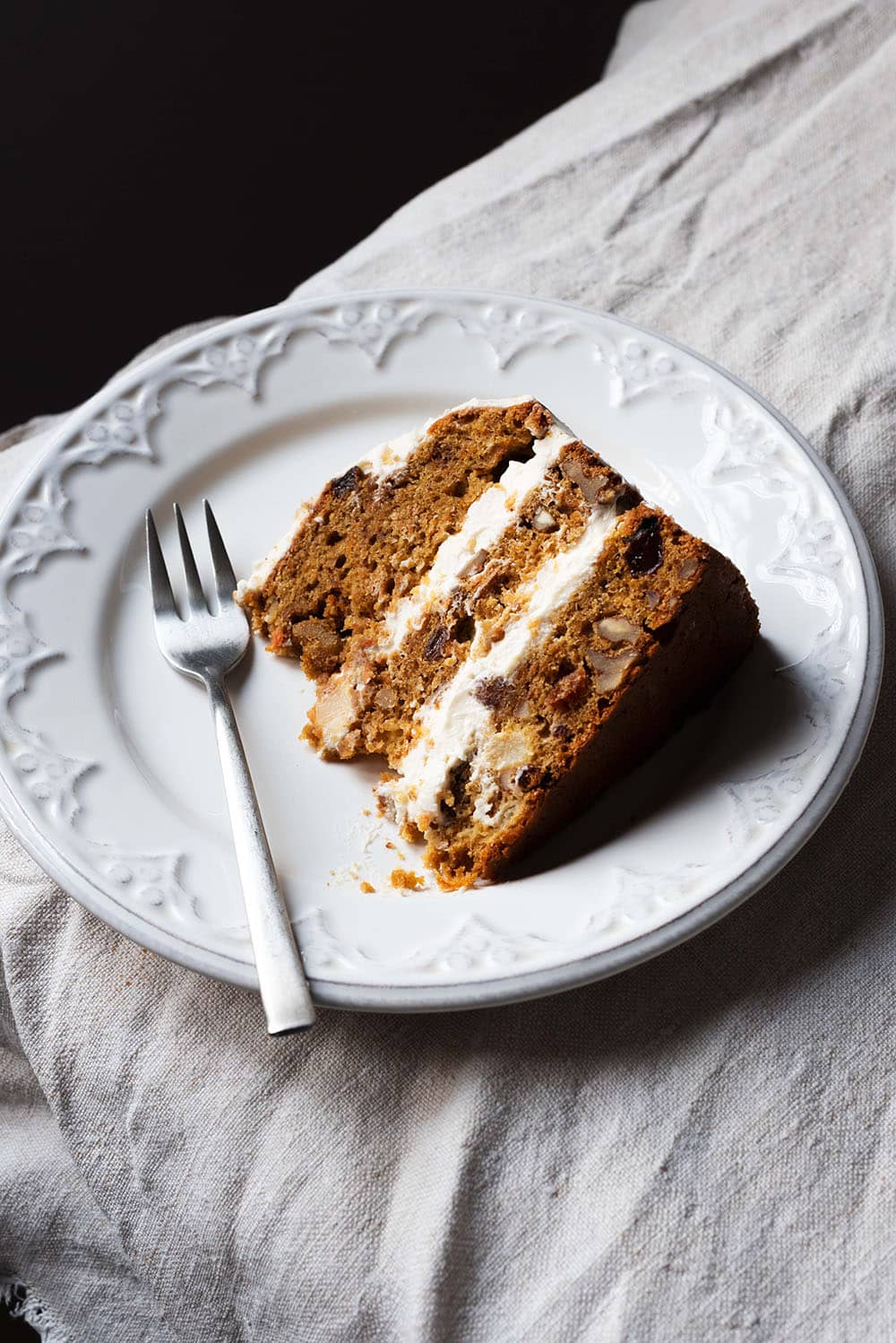 Hay un mantel blanco en la mesa.Encima hay un plato blanco. Encima hay una ración de Carrot Cake. Se ve bien y gustoso sus capas.