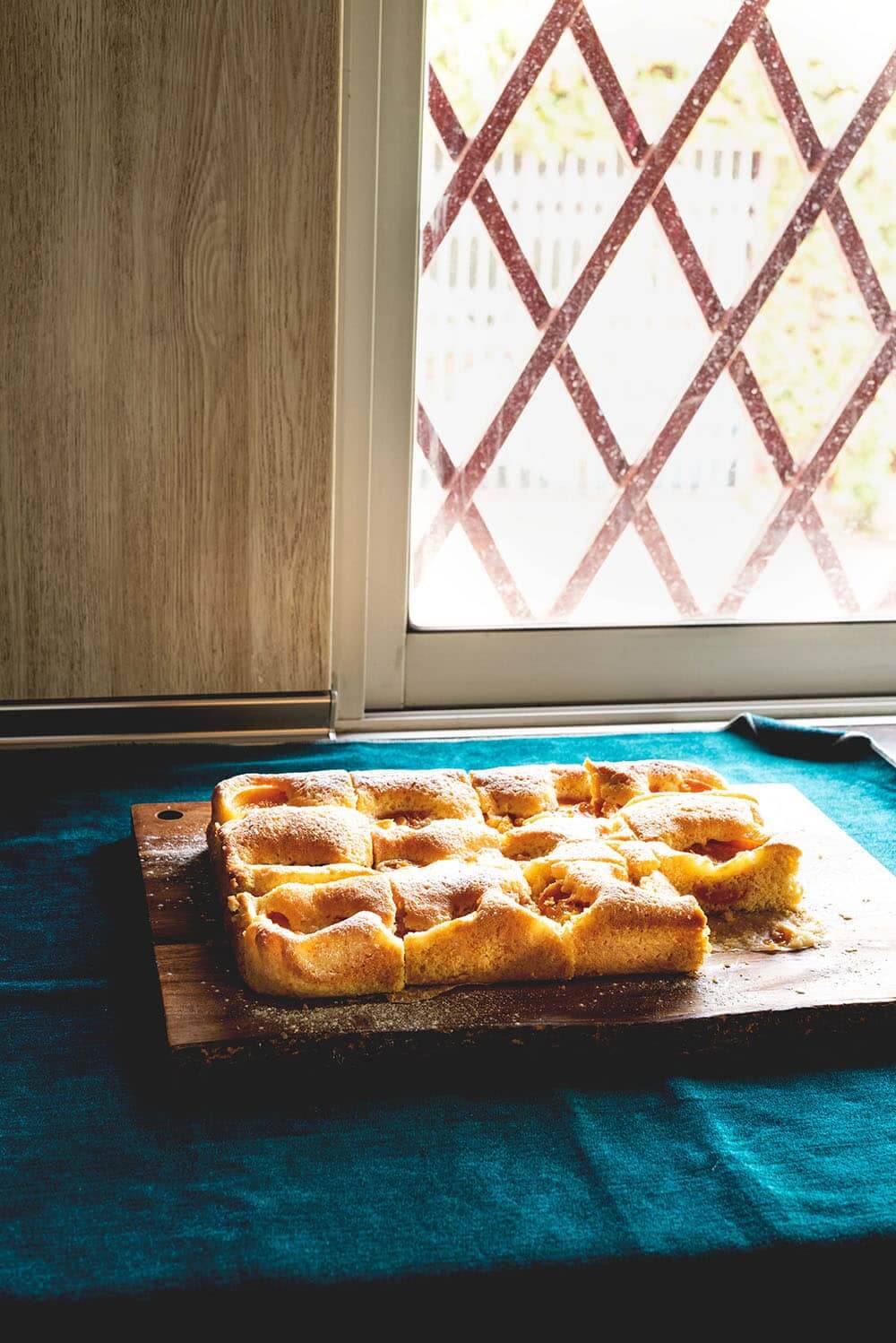 En una encimera hay un mantel azul. Encima está el Marillenkuchen cortado. Se ve esponjoso.