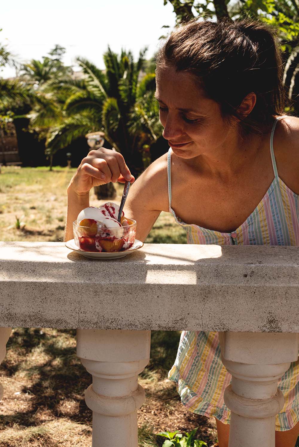 Una persona está catando en el jardín una ración de Peach Melba. El postre está en un vaso de vidrio. Este recipiente está encima de una valla de piedra. 