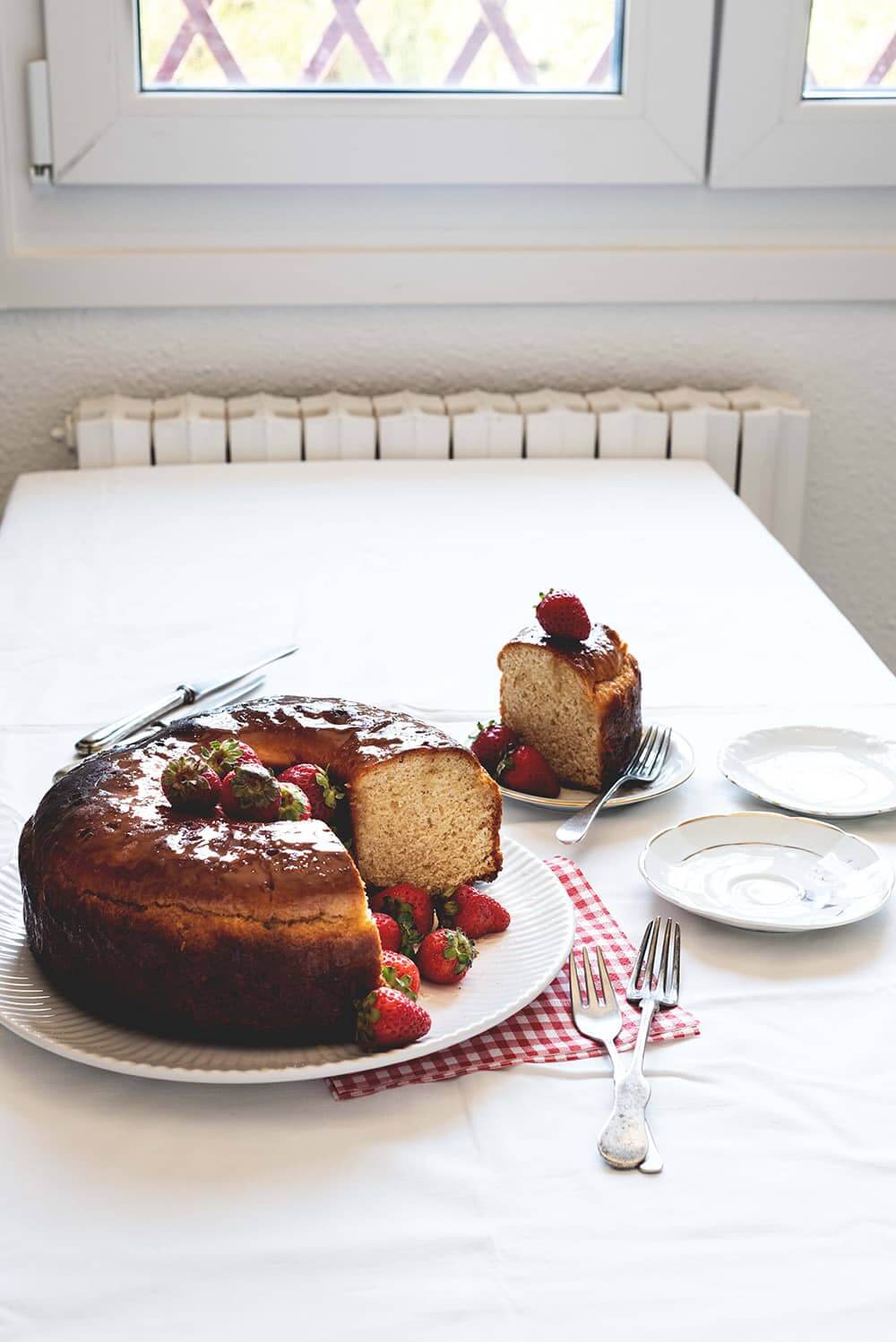 Receta de Savarin de fresas Hay un mantel blanco en la mesa.Encima hay un plato blanco. Encima está el Savarin francés con fresas ya cortado. Alrededor del postre hay cuchillos y platos blanco, pala de tarta y un plato con una ración de Savarin.