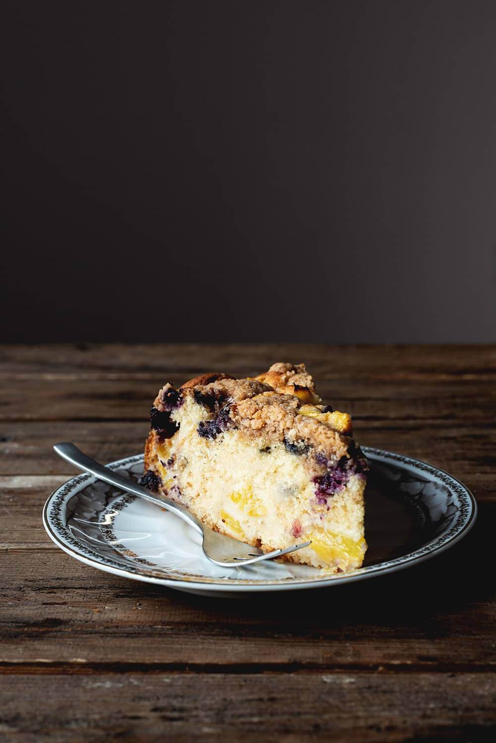 Receta de coffee cake de arándanos y melocotón En una mesa madera hay un plato blanco y marrón. Encima hay una ración de coffee cake de arándanos y melocotón. Se ve esponjoso y jugoso por las frutas. Al lado del postre hay un tenedor.