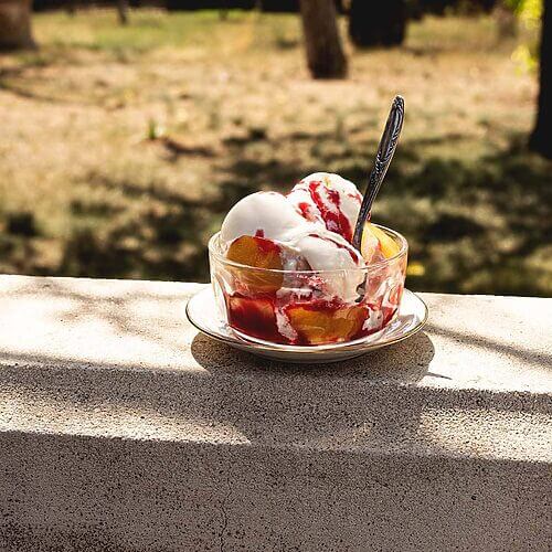 En una valla de piedra hay un plato blanco y encima hay un recipiente transparente. Dentro está el helado Peach Melba: helado de vainilla, salsa de frambuesa y melocotones frescos.