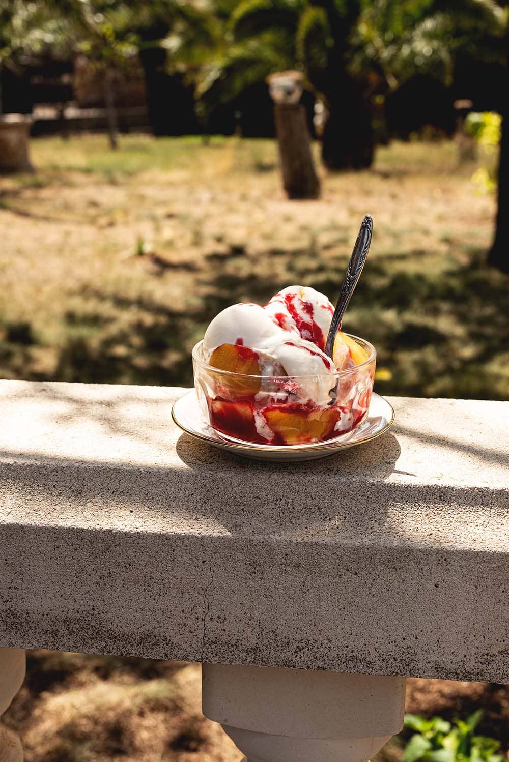 En una valla de piedra hay un plato blanco y encima hay un recipiente transparente. Dentro está el helado Peach Melba: helado de vainilla, salsa de frambuesa y melocotones frescos.