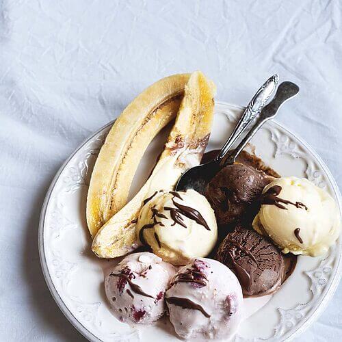 En una mesa hay un mantel blanco. Encima hay un plato blanco. Encima está el postre Banan Split: helado de vainilla, de fresa y de chocolate. Todo acompañado con salsa de chocolate y plátano cortado en la mitad.