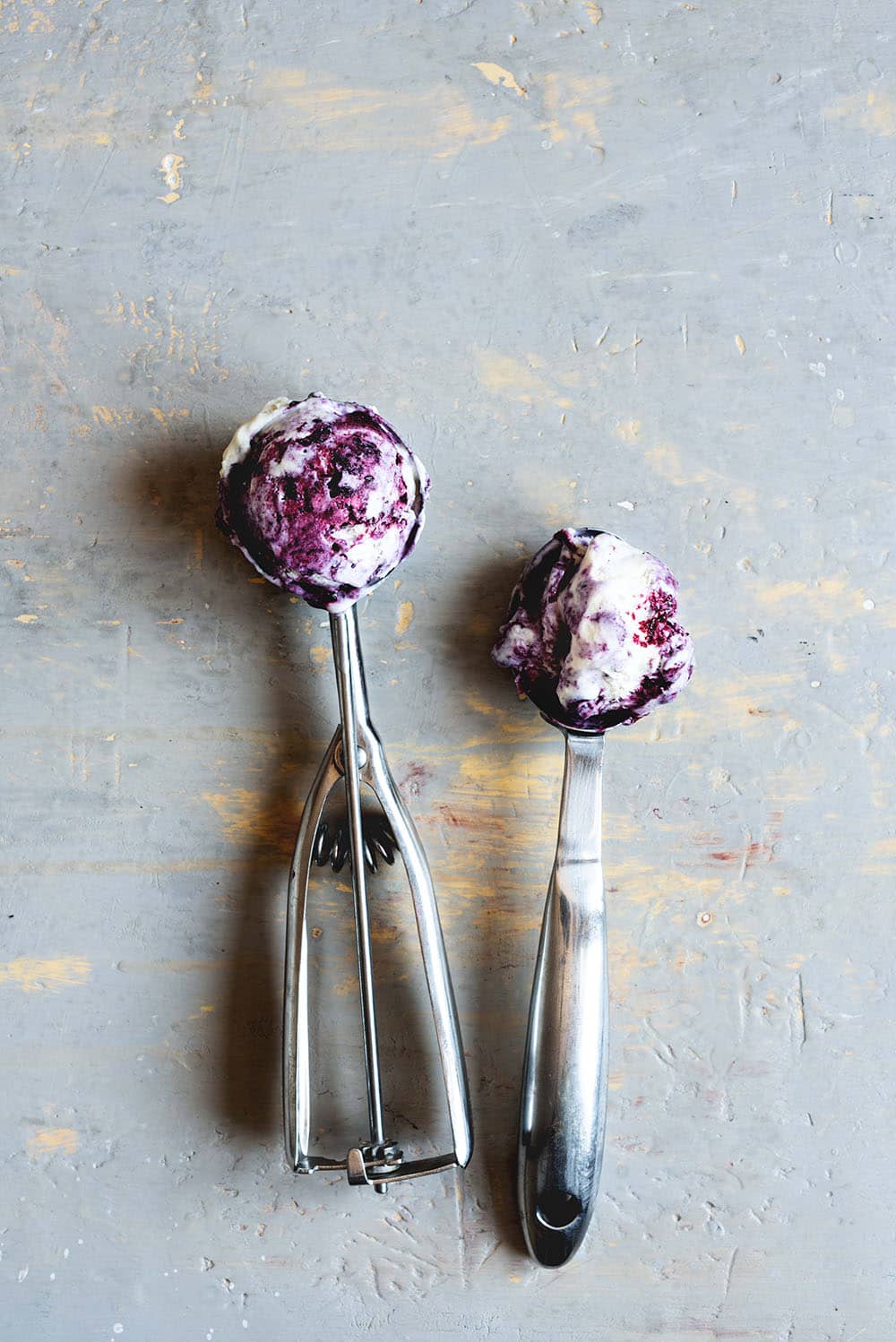 Receta de helado de frutos rojos En una mesa gris hay dos sacabolas de helado lleno de helado de frutos rojos. Se ven cremosos.