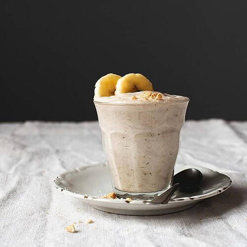 En una mesa hay un mantel blanco. Encima hay un plato blanco . Encima hay un vaso transparente con mousse de plátano. Está decorada con dos rodajas de plátanos. Al lado del vaso hay una cuchara y virutas de galletas.