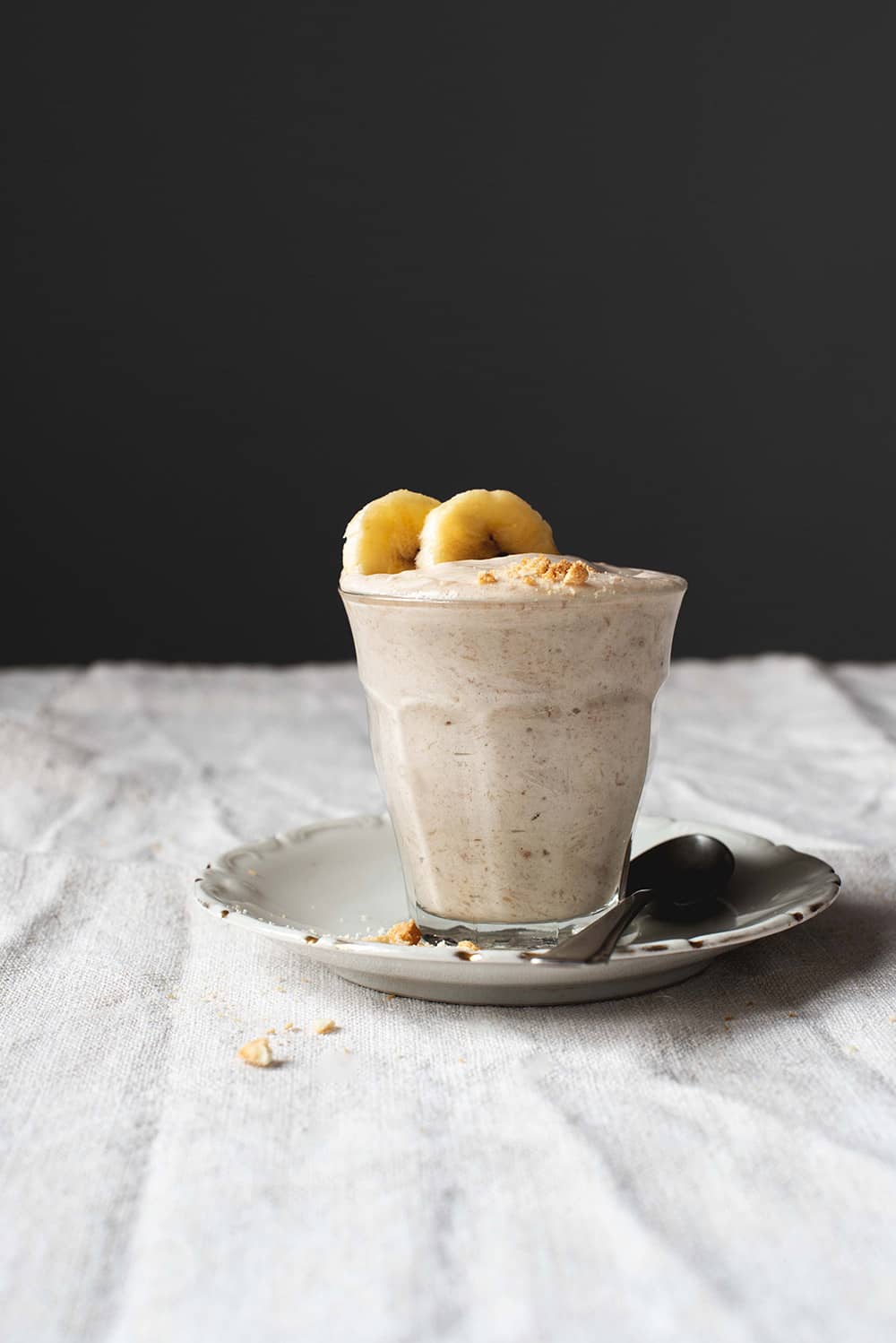 En una mesa hay un mantel blanco. Encima hay un plato blanco . Encima hay un vaso transparente con mousse de plátano. Está decorada con dos rodajas de plátanos. Al lado del vaso hay una cuchara y virutas de galletas.