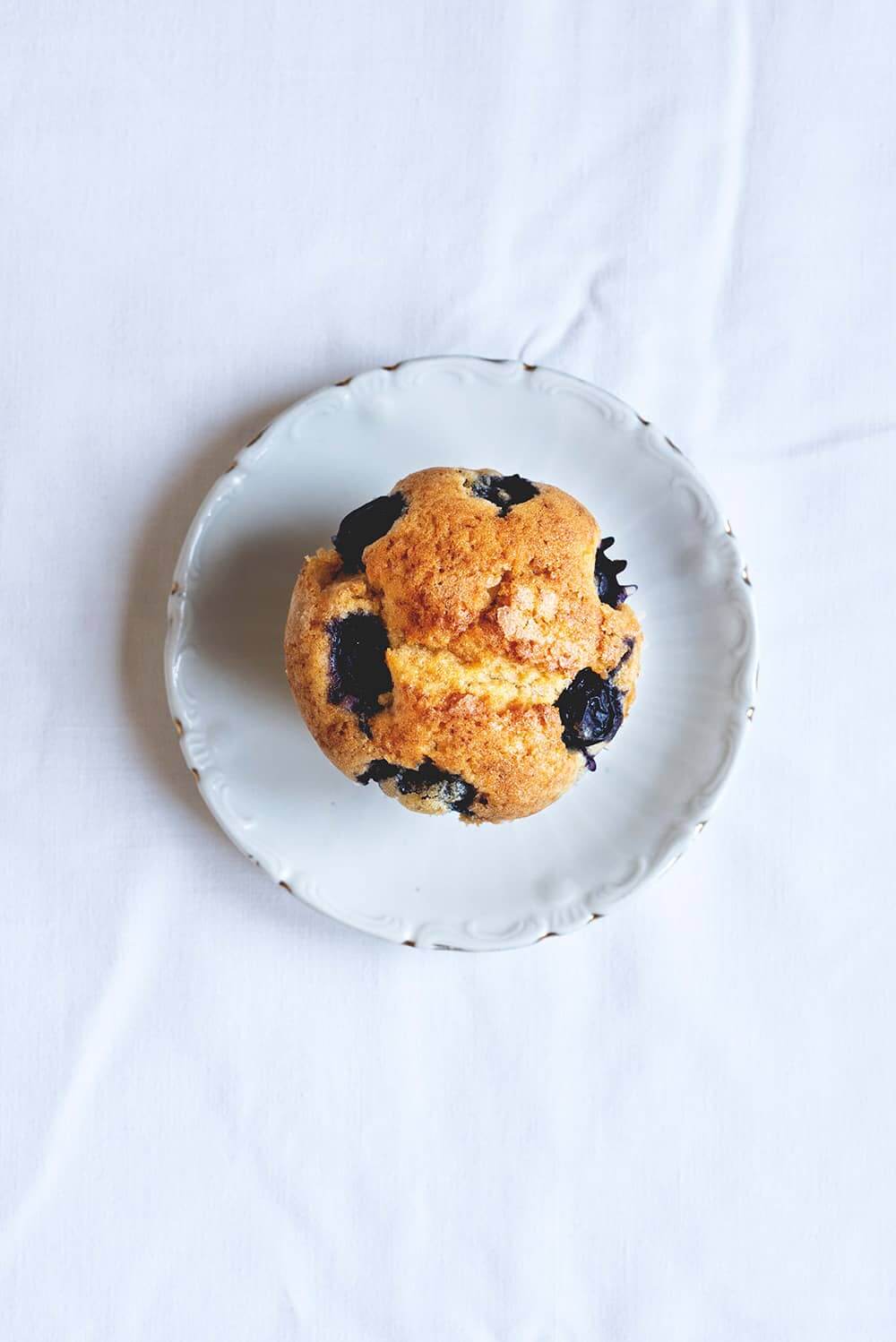 En una mesa de madera hay un mantel blanco. Encima hay un plato blanco encima está el muffin de arándanos. La foto es cenital.