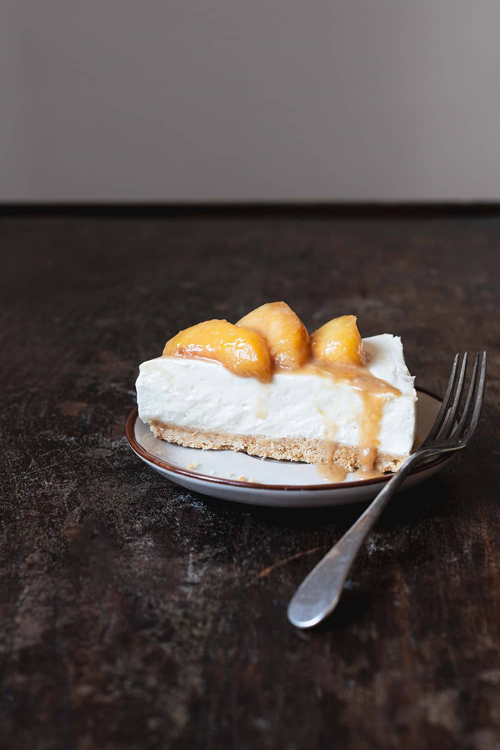 Receta de tarta de queso y melocotón sin horno En una mesa de madera hay un plato marrón claro. Encima está una ración de tarta de queso y melocotón sin horno. Se ven los melocotones salteados jugosos y la tarta de queso cremoso. Al lado del postre hay un tenedor.