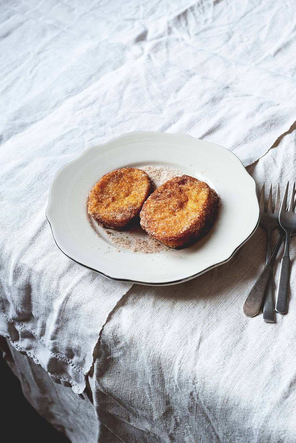Hay un mantel blanco en la mesa. Encima hay un plato blanco. Encima del plato hay dos torrijas de leche . Una está cortada. Se ve su interior cremoso.