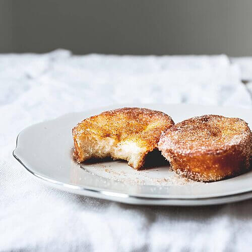 Hay un mantel blanco en la mesa. Encima hay un plato blanco. Encima del plato hay dos torrijas de leche. Están doradas y apetitosas.