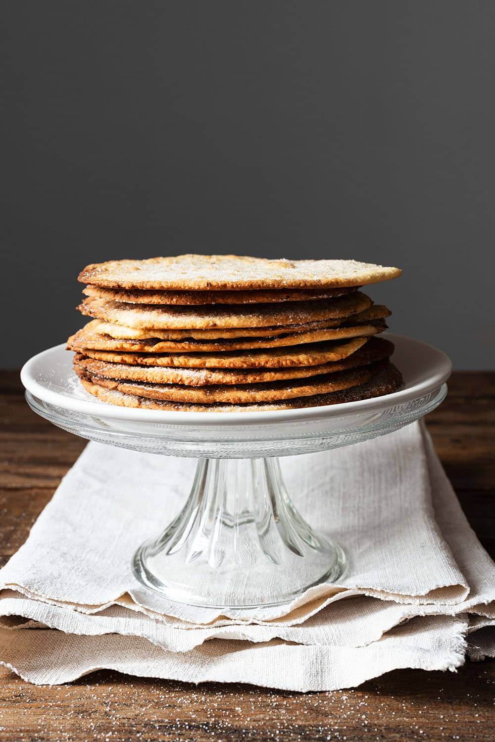 Receta de tortas de anís y aceite crujientes En una mesa de madera hay un paño blanco. Encima está una portatarta de cristal. Encima están amontonados las tortas de anís y aceite crujientes.