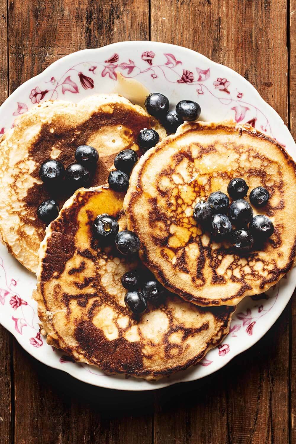 Es una foto cenital. Se ve una mesa de madera. Encima está un plato blanco vintage con las tortitas americanas bañadas con miel y acompañadas con arándanos frescos.