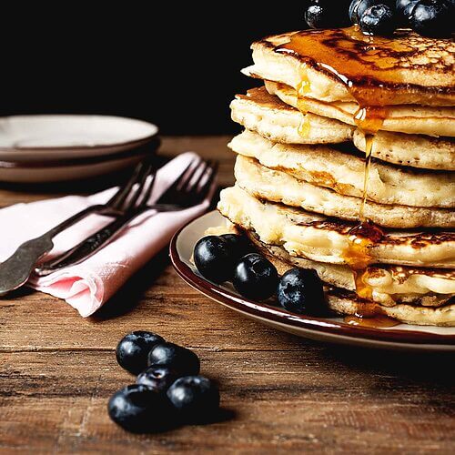 En una mesa de madera hay un plato blanco. Encima hay muchas tortitas americanas esponjosas. Están bañadas con miel y acompañadas con arándanos. Alrededor del plato hay una servilleta rosa tenedores y arándanos.
