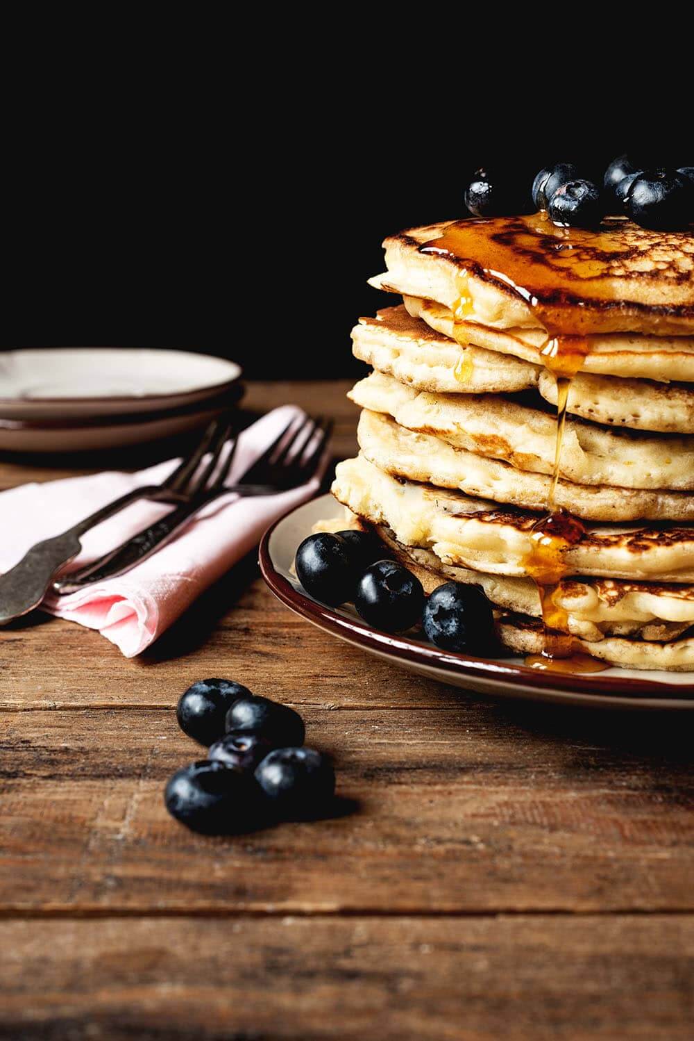 En una mesa de madera hay un plato blanco. Encima hay muchas tortitas americanas esponjosas. Están bañadas con miel y acompañadas con arándanos. Alrededor del plato hay una servilleta rosa tenedores y arándanos.