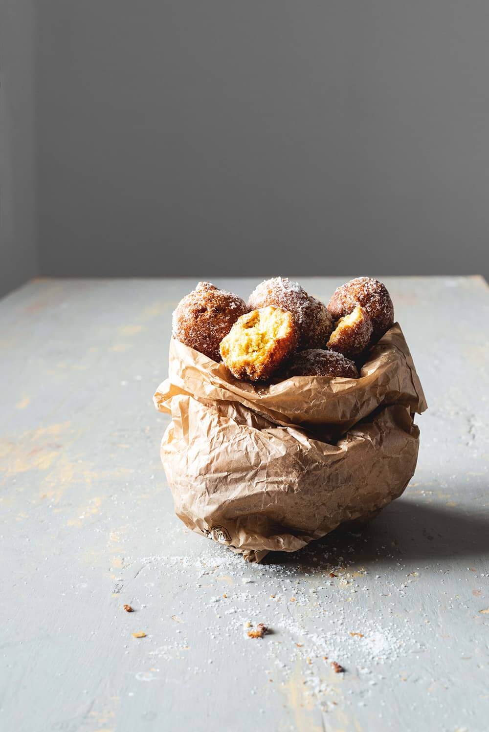 En una mesa gris hay una bolsa de papel rústico llena de Castagnole. Un buñuelo está cortado y se ve esponjoso. Alrededor de la bolsa hay azúcar y migas.