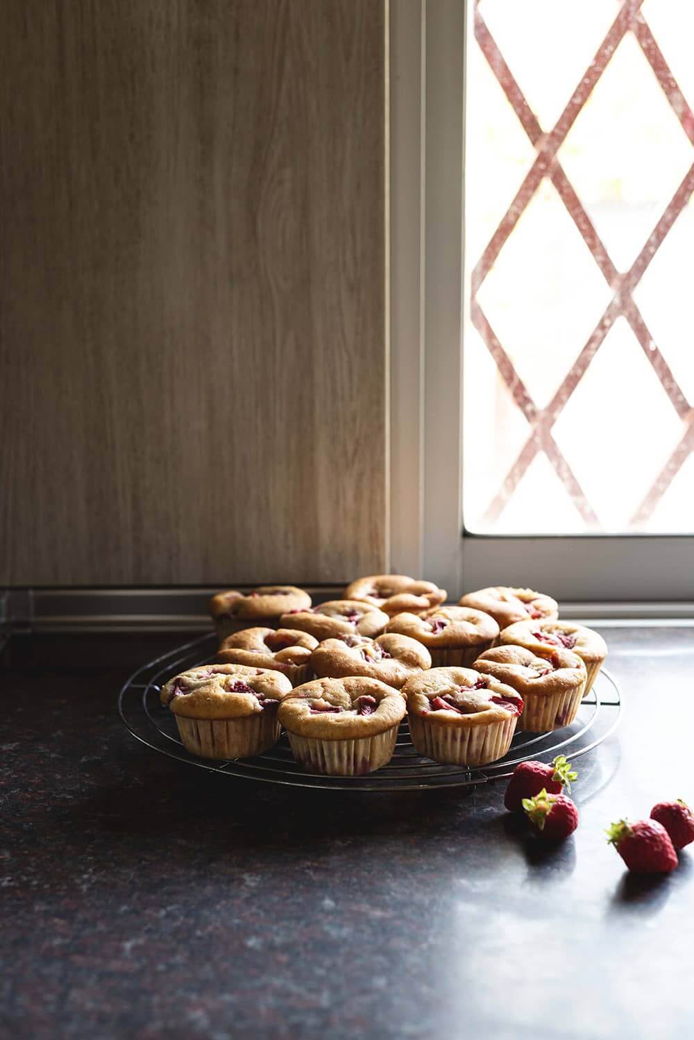 Receta magdalenas con fresas En una encimera gris hay una rejilla. En la rejilla están las magdalenas con fresas enfriándose. Alrededor de las magdalenas hay un par de fresas.