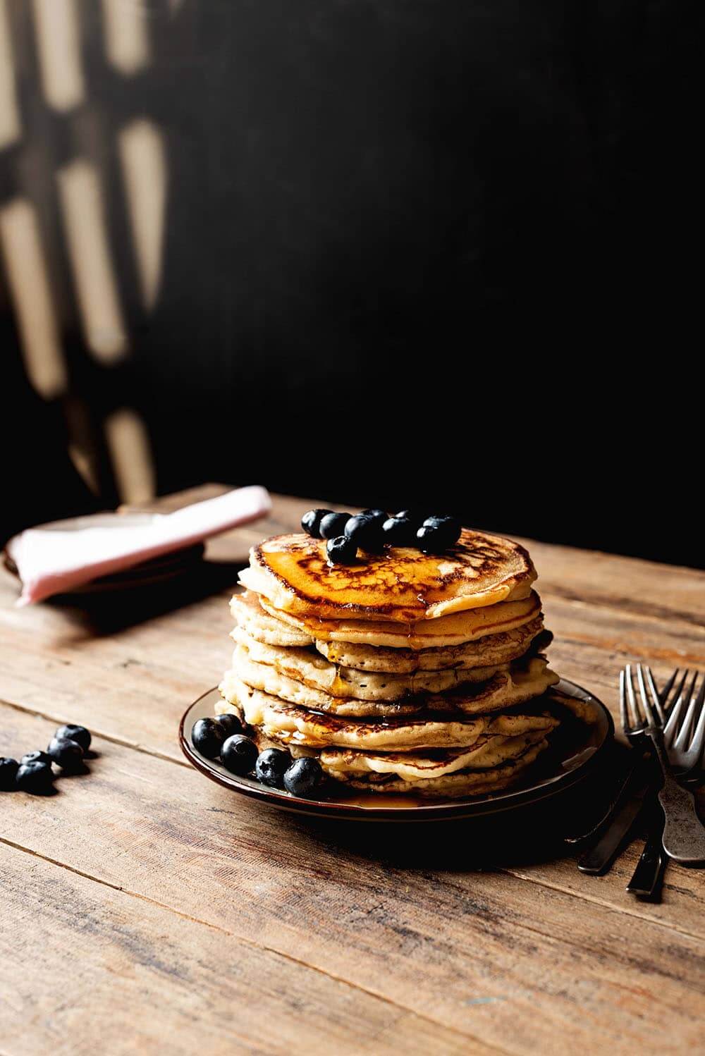 En una mesa de madera hay un plato blanco. Encima hay muchas tortitas americanas esponjosas. Están bañadas con miel y acompañadas con arándanos. Se ven sombras bonitas en la pared. En la mesa además hay una servilleta de tela de color rosa, platos y arandnos.