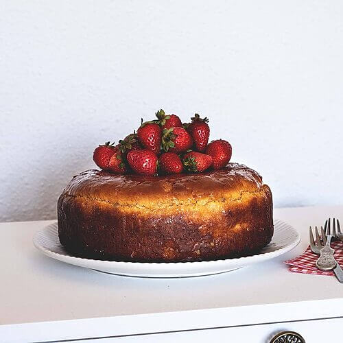 En una cómoda blanca está Savarin al ron y fresas. Al lado del plato, que alberga el postre, hay una servilleta roja, Encima hay un par de tenedores.