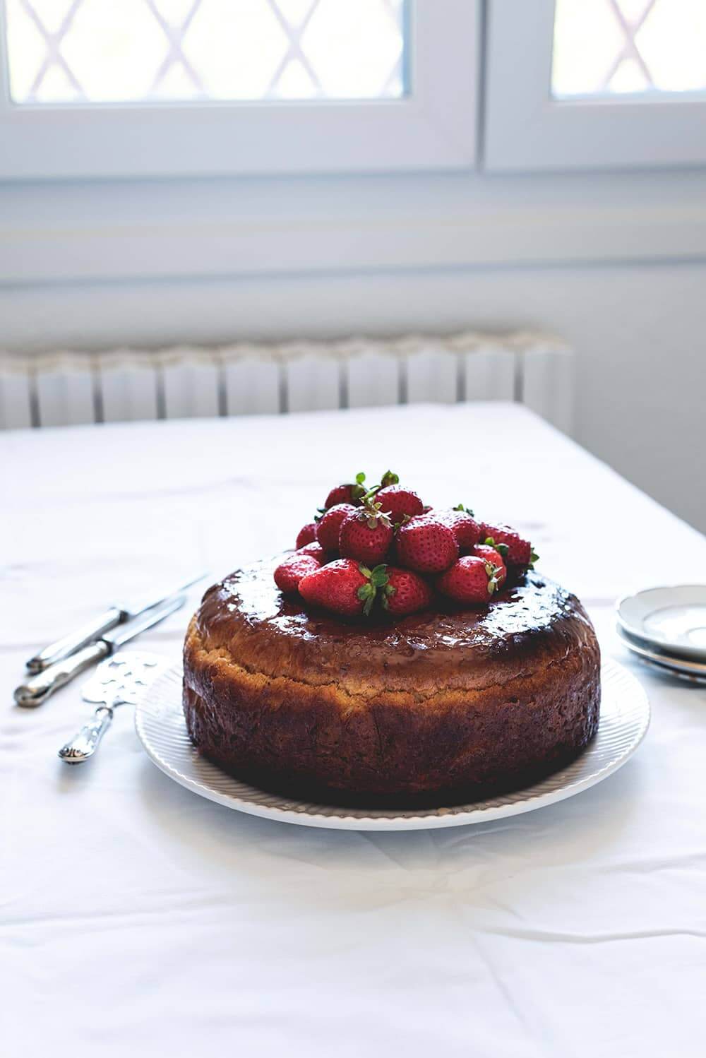 Savarin francés con fresas Hay un mantel blanco en la mesa.Encima hay un plato blanco. Encima está el Savarin francés con fresas ya cortado. Alrededor del postre hay cuchillos y platos blanco, pala de tarta y un plato con una ración de Savarin.