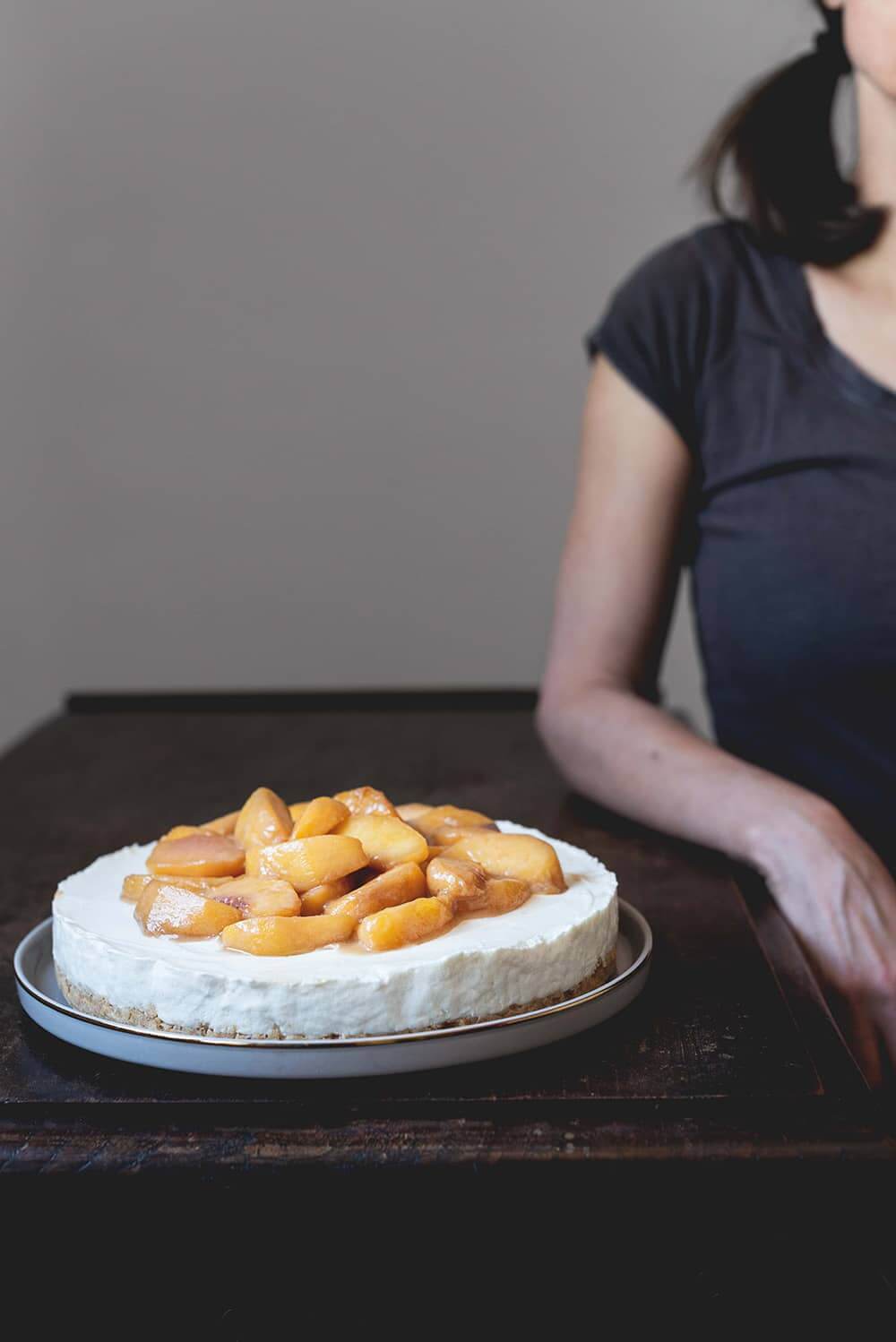 Tarta de melocotón con queso philadelphia En una mesa de madera hay un plato blanco. Encima está tarta de melocotón con queso philadelphia. Al lado del postre hay una persona esperando para poder cortarlo.