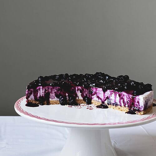 En una mesa de madera hay un mantel blanco. Encima está la portatarta blanca y encima de ella está la tarta de queso con arándanos philadelphia sin horno. Ya está cortada. Se ve jugoso y empapado por la salsa de arándanos.