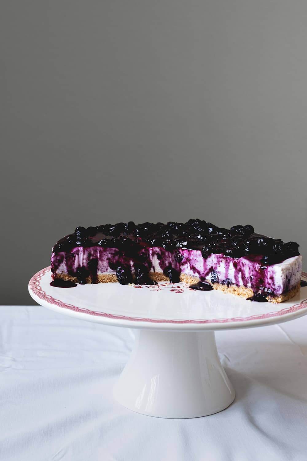En una mesa de madera hay un mantel blanco. Encima está la portatarta blanca y encima de ella está la tarta de queso con arándanos philadelphia sin horno. Ya está cortada. Se ve jugoso y empapado por la salsa de arándanos.
