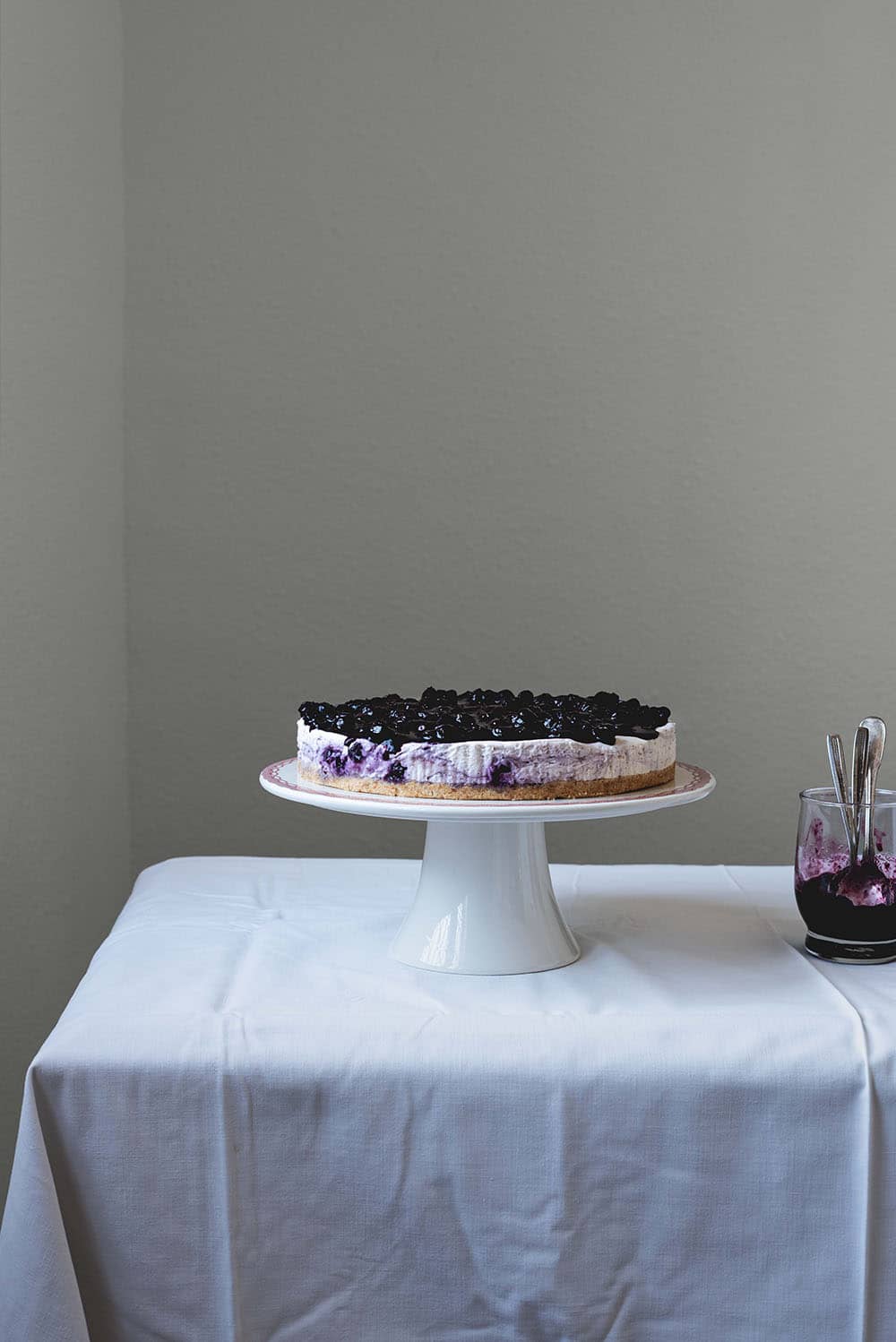 En una mesa de madera hay un mantel blanco. Encima está la portatarta blanca y encima de ella está la tarta de queso con arándanos y base de galleta sin horno. Al lado está un vaso con salsa de arándanos.