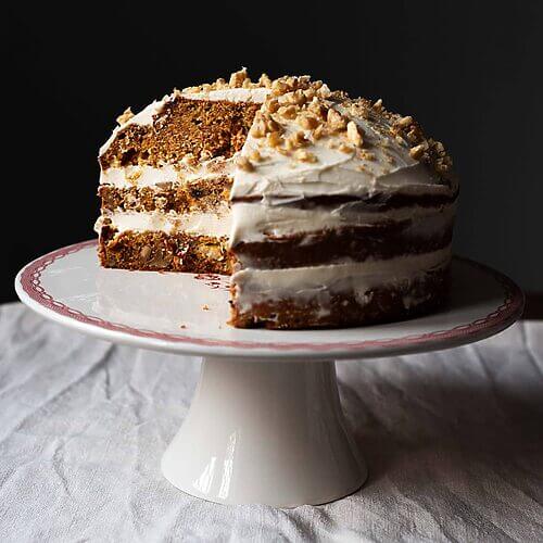 Hay un mantel blanco en la mesa. Encima hay una portatarta blanca. Encima de ella está la tarta de zanahoria americano cortada. 
