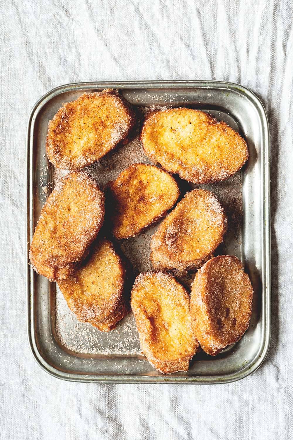 Hay un mantel blanco en la mesa. Encima hay una bandeja de hojalata con varias torrijas de leche tradicionales. Se ven gustosas y doradas.