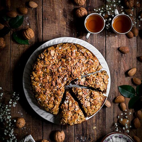 En una mesa de madera hay un plato blanco. Encima está el Bizcocho de nueces y almendras. ya está cortada. Se ve muy delicioso. Alrededor hay tazas de té llenas de té, flores, almendras, cubiertos y un paño de cocina blanca.
