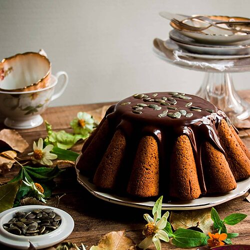 En una mesa de madera hay un plato blanco. Encima está el Bundt cake de calabaza con salsa de chocolate. El chocolate es brillante y muy apetecible. Alrededor del postre hay platos, flores, tazas, hojas y flores.