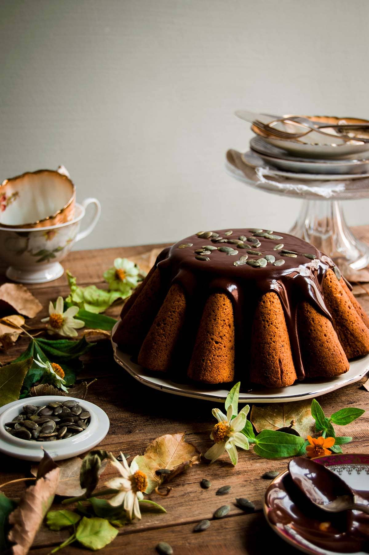 En una mesa de madera hay un plato blanco. Encima está el Bundt cake de calabaza con salsa de chocolate. El chocolate es brillante y muy apetecible. Alrededor del postre hay platos, flores, tazas, hojas y flores.