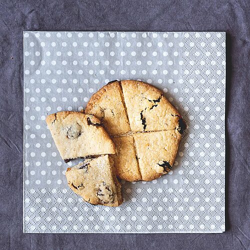 En una servilleta gris con lunares blancos están dos galletas de Soul Cake . Una está cortada y se ve crujiente y se aprecian las pasas y su corte original.