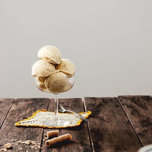 En una mesa de madera hay una copa llena de helado casero de vainilla sin máquina. Alrededor hay galletas desmigadas.