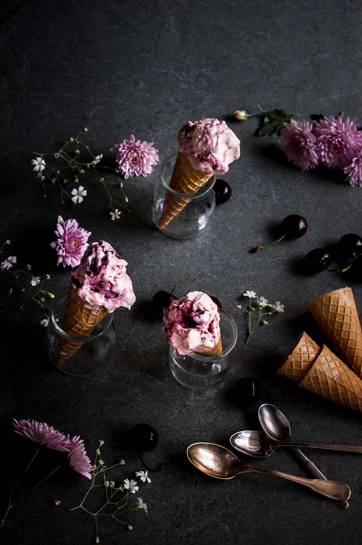 En una mesa de piedra hay tres vasos de cristal. En cada una hay cucuruchos y helado de cheesecake con cerezas sin heladera. Alrededor hay cerezas, flores y cucuruchos vacíos.