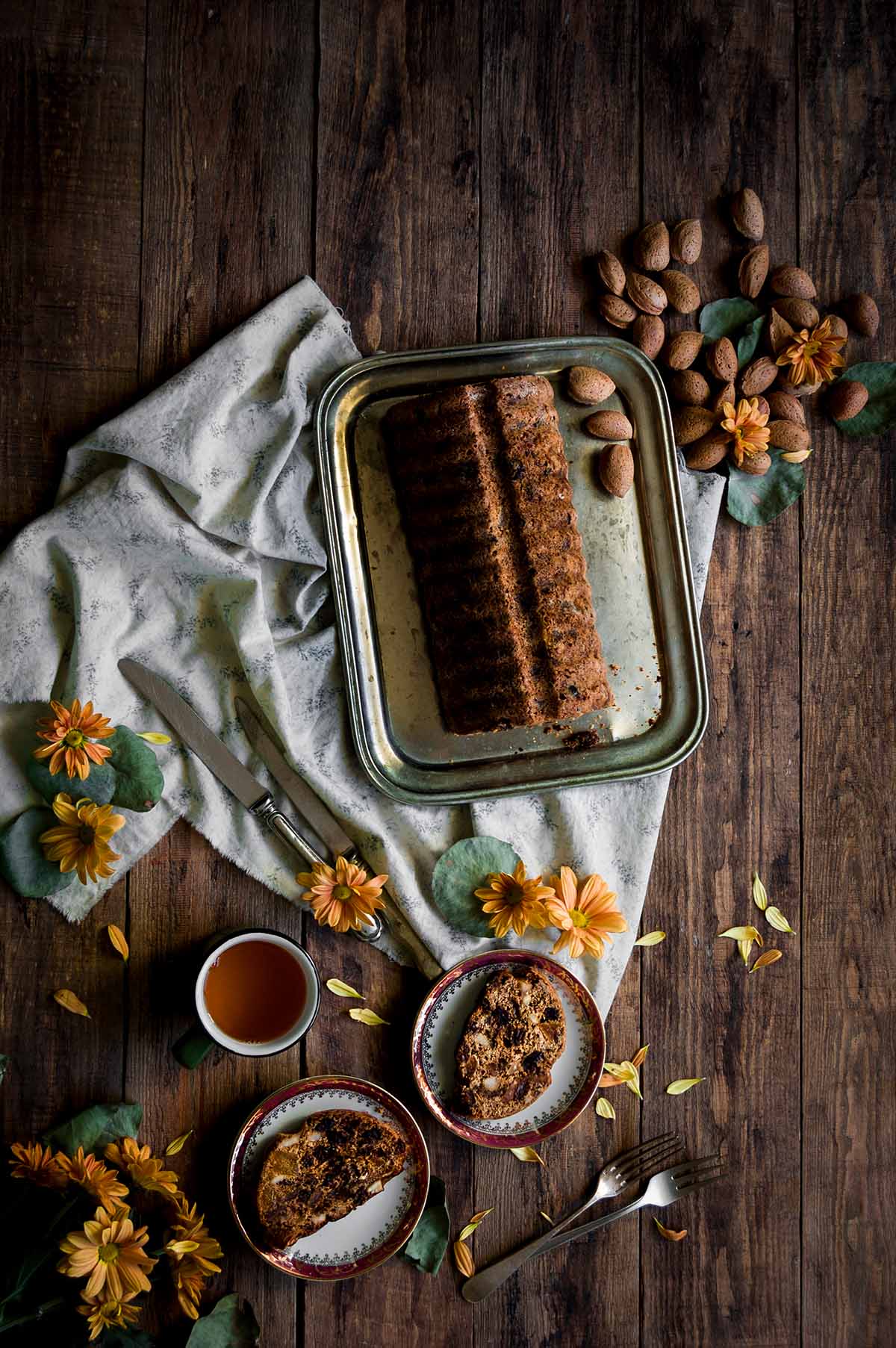 En una mesa de madera está puesto un paño de cocina blanca Encima está una bandeja rectangular metálica. Encima de esta bandeja está el pan de frutas. Está cortado. Se ve muy jugoso y delicioso por las frutas secas. Alrededor hay platos con el postre, flores y cubiertos.