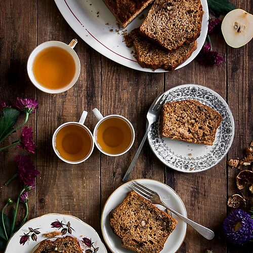 En una mesa de madera está puesto un plato ovalado blanco. Encima está el plum cake de peras esponjoso. ya está cortado.Se ve su interior esponjoso y tierno. Alrededor del postre hay platos, cubiertos, tazas de té y flores.